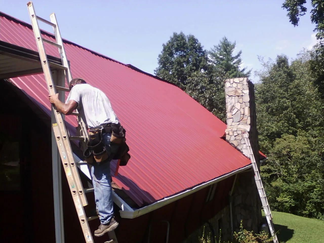 Roofer on a ladder beside a red metal roof and stone chimney — Soffit & Fascia Repair by Roofing Brothers in Salamanca